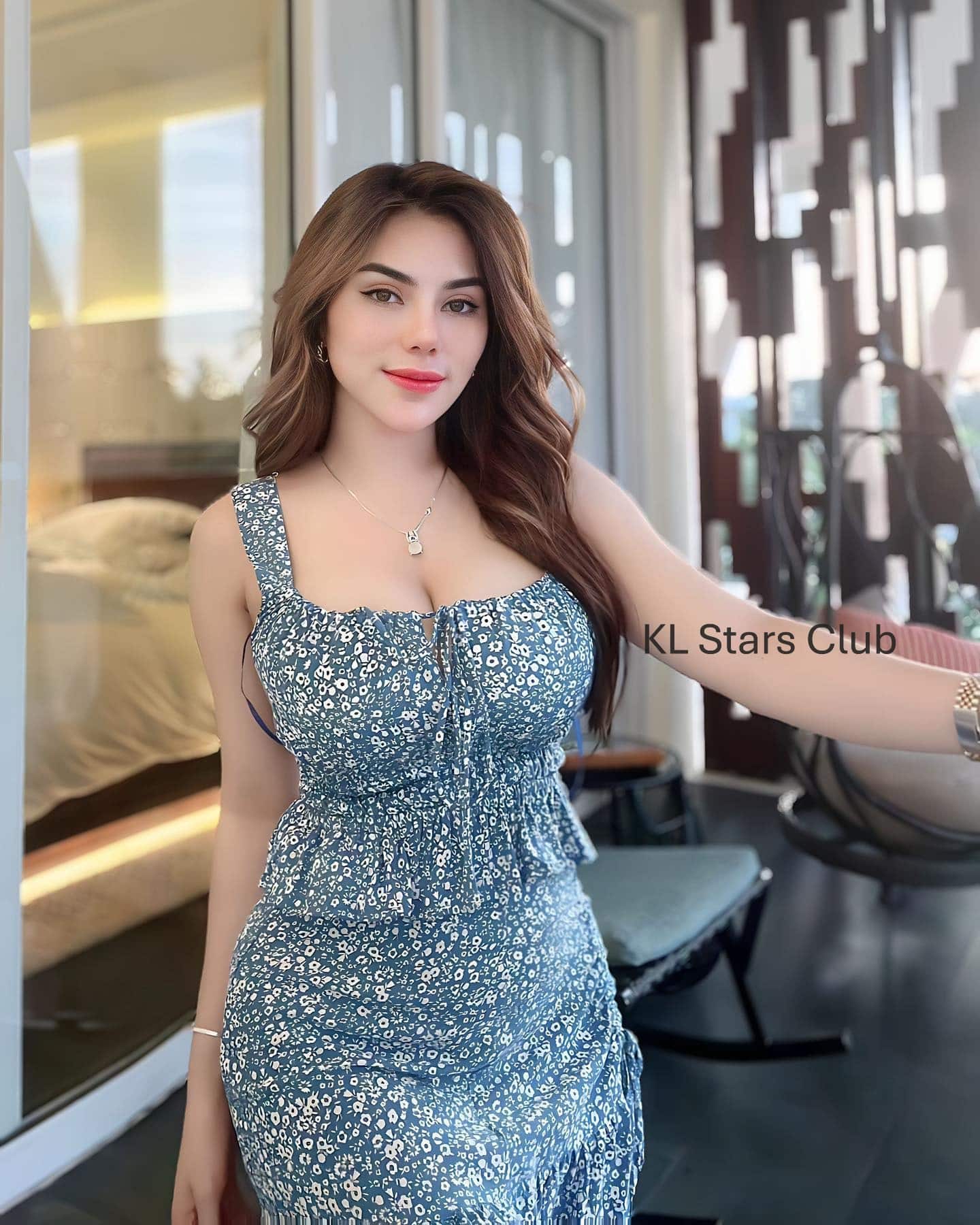 Kuala Lumpur escort Victoria Qistina in a blue floral dress, posing on a modern balcony with glass doors in the background.